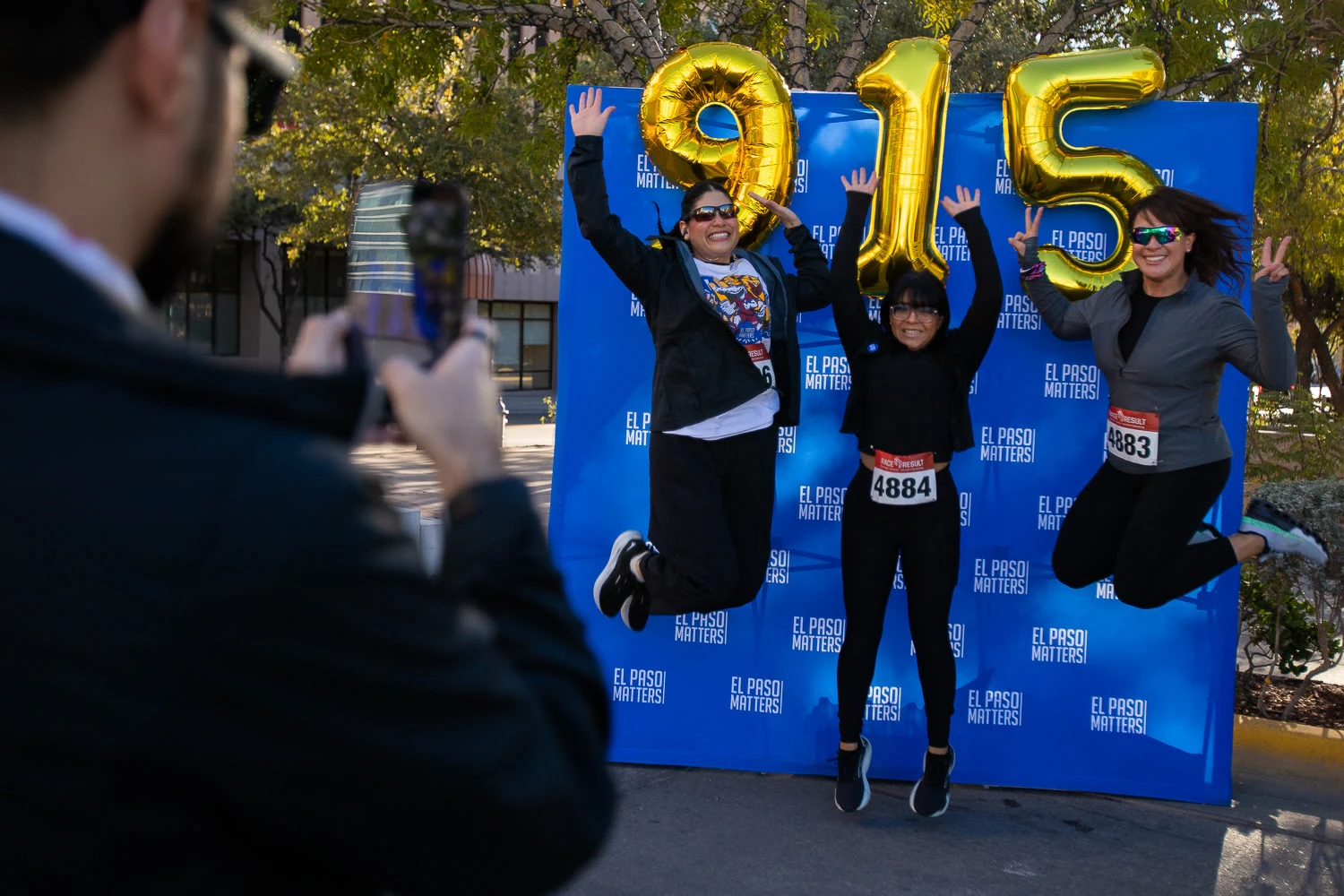 9.15K runners jump for joy in front of an El Paso Matters banner and 9-1-5 balloons